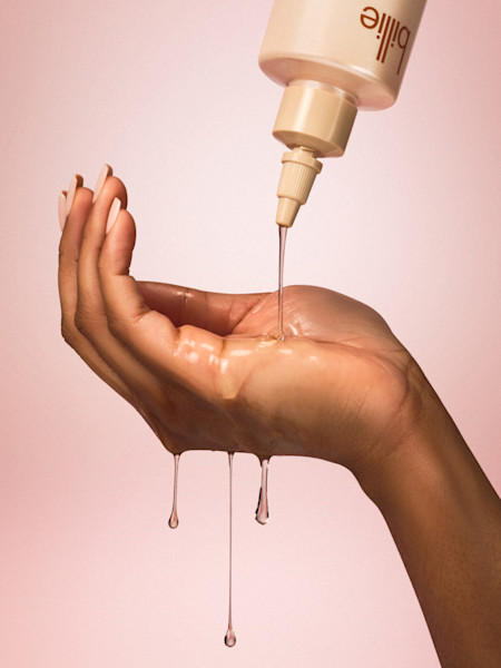 Clear skincare product being dispensed onto a hand with drops dripping down against a soft pink background.
