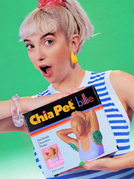 Person with blonde hair and yellow earrings holding a Chia Pet Billie product against bright green background.