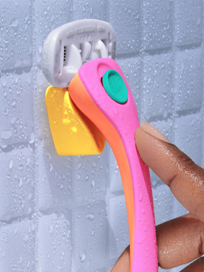 Hand removing a pink and orange Billie razor from a yellow magnetic holder on a white tiled shower wall
