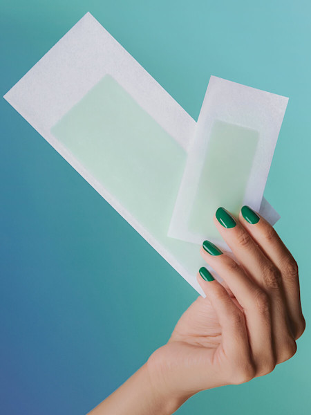 Hand with green nail polish holding two different size body and underarm wax strips against a blue and green gradient background