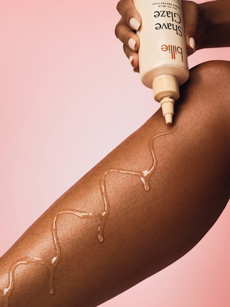 Hand with white nails applying Billie Shave Glaze product in a zigzag pattern on skin against pink background.